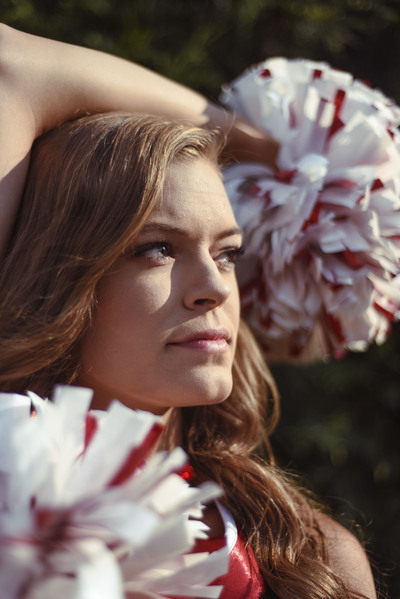 a high school senior poses in their cheer outfit with their pom poms framing their face staring into the distance