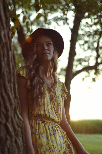 a young lady leans agains a tree with the sunset behind her