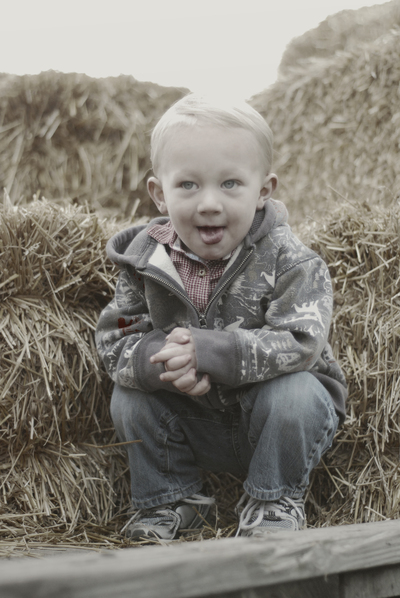 a young boy squats on a pile of hay with their tongue sticking out