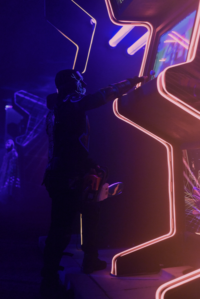 a scare actor in a mask with a chainsaw gets distracted playing an oversized neon arcade game