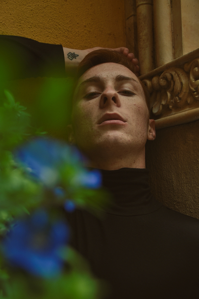 a young adult sits against a wall hiding behind blue flowers